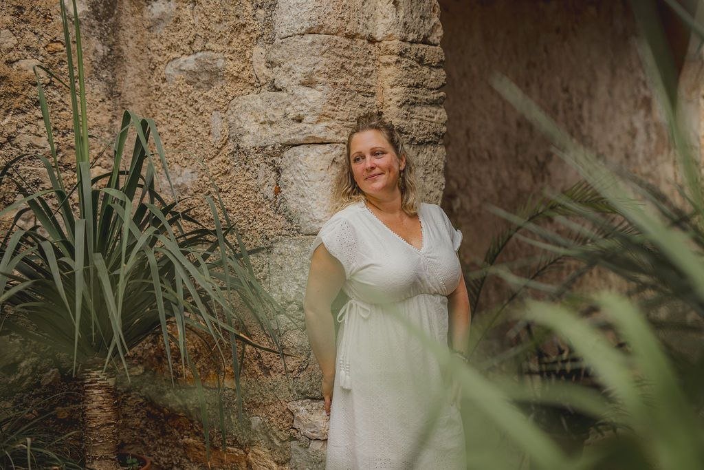 Sarah im weißen Kleid vor einer Natursteinwand, umgeben von Pflanzen.