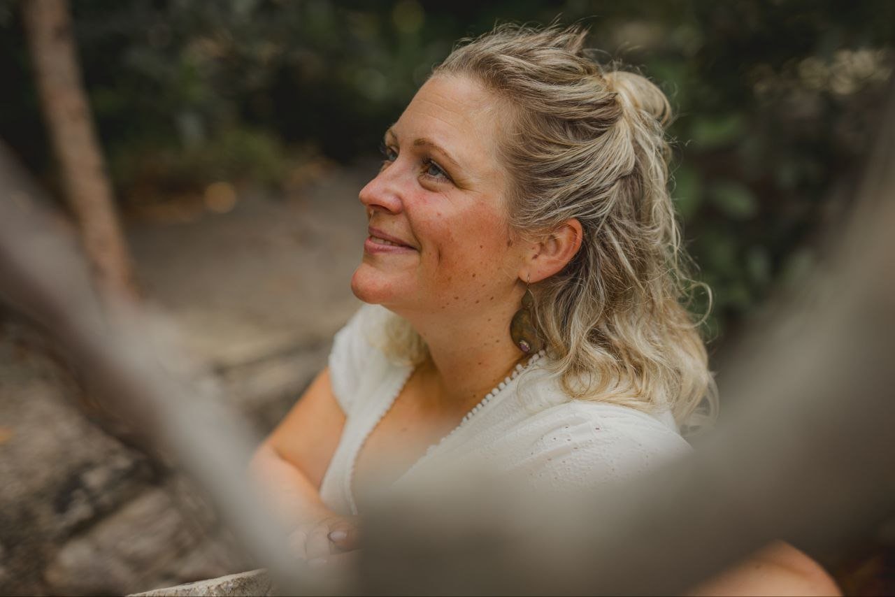 Sarah im Porträt zwischen Bäumen und warmen Natursteinen.